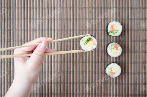 A hand with chopsticks holds a sushi roll on a bamboo straw serwing mat background